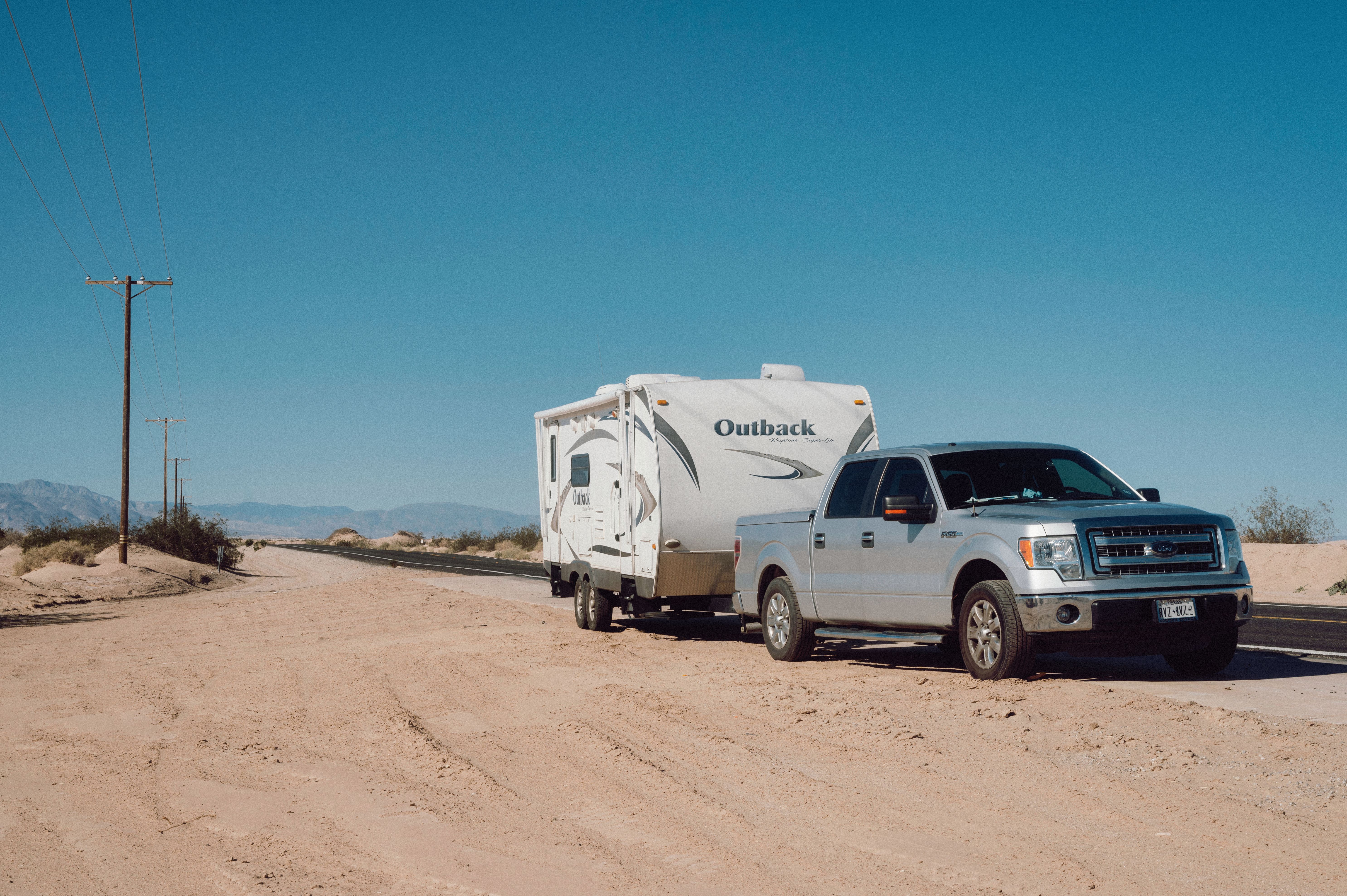 Silver pickup truck towing a travel trailer along a desert road under a clear sky.
